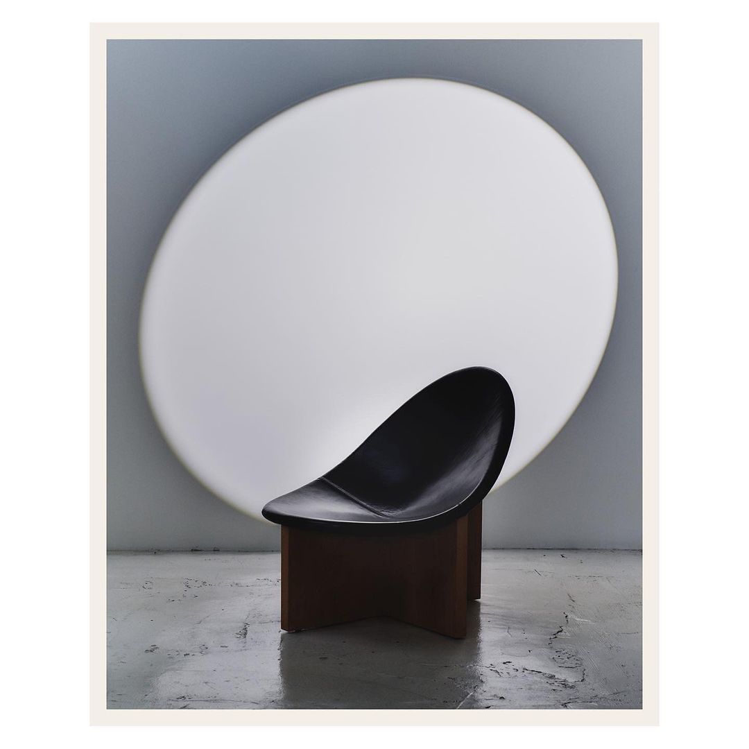 Modern black chair with a brown base, elegantly posed against a large, round, softly illuminated white backdrop. Studio lighting.