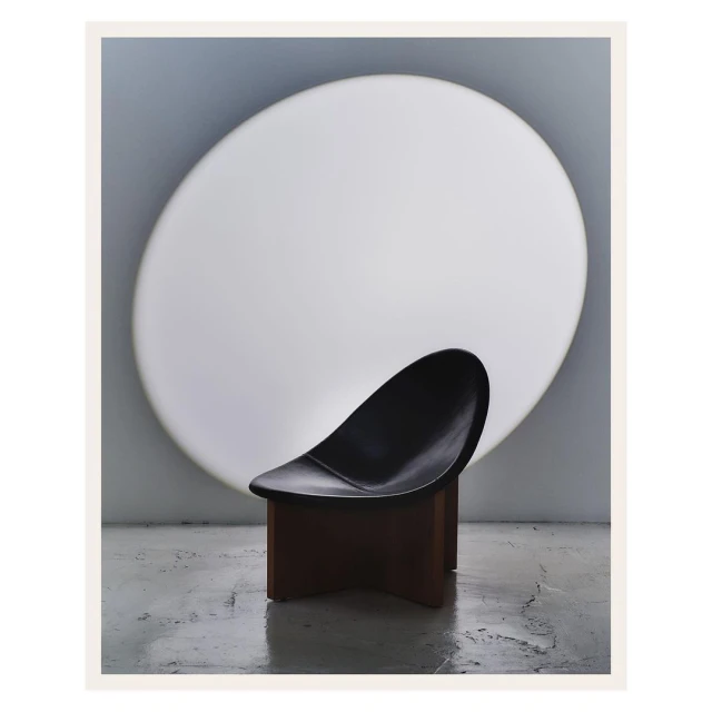 Modern black chair with a brown base, elegantly posed against a large, round, softly illuminated white backdrop. Studio lighting.