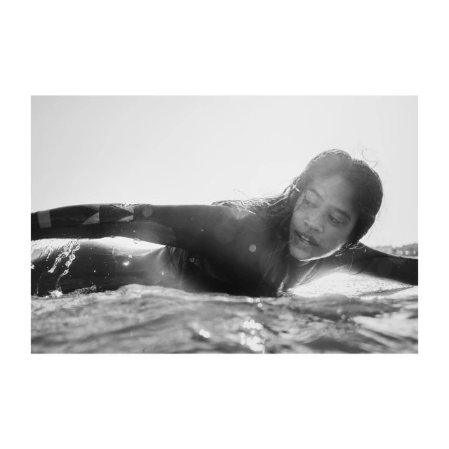 Black and white shot of a person lying on a surfboard in the ocean, with water droplets visible and bright sunlight overhead.