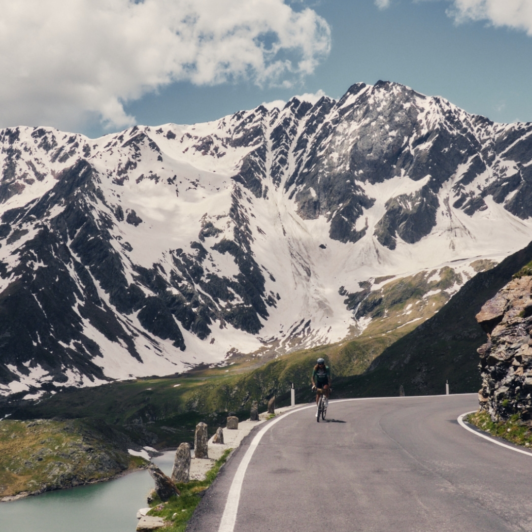 Cyclist riding uphill on a mountain road, with a lake and snow-capped peaks in the background on a sunny day under a cloudy sky.
