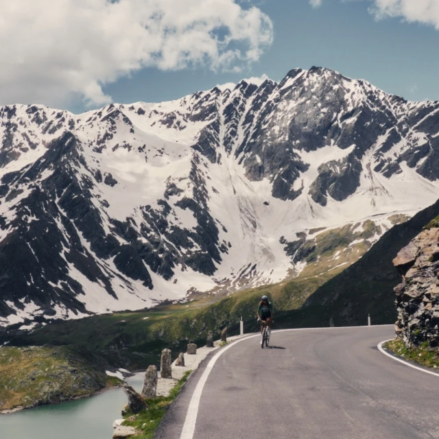 Cyclist riding uphill on a mountain road, with a lake and snow-capped peaks in the background on a sunny day under a cloudy sky.