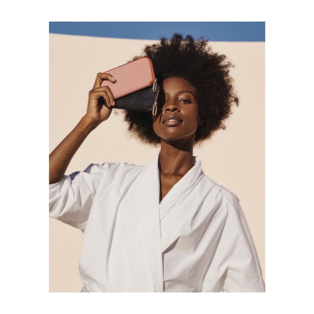 A woman with afro hair holds a pink and navy wallet against a light beige background, wearing a white robe-like shirt.