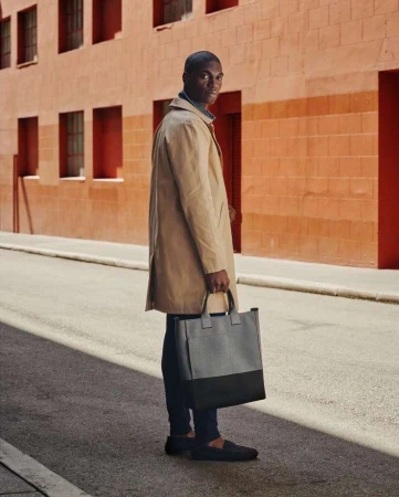 Man wearing a beige trench coat holding a two-tone grey tote bag while standing on an asphalt street