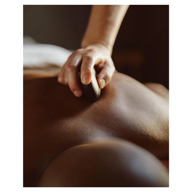 Adult receiving a hot stone massage on their back, hand with stone pressing into the skin, spa treatment, dark background.