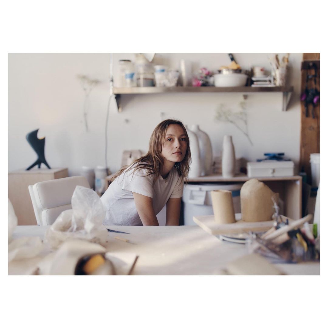 Woman in white t-shirt poses in art studio filled with clay and ceramic art, lit by natural light with a shallow depth of field.