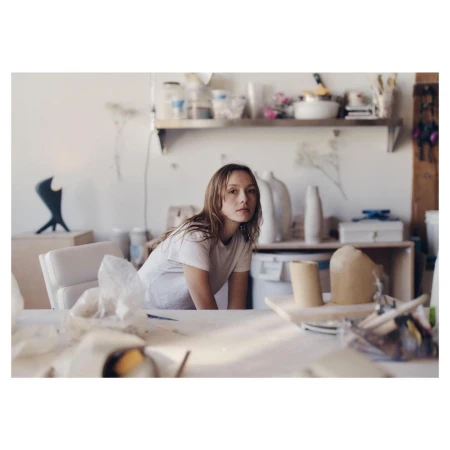 Woman in white t-shirt poses in art studio filled with clay and ceramic art, lit by natural light with a shallow depth of field.
