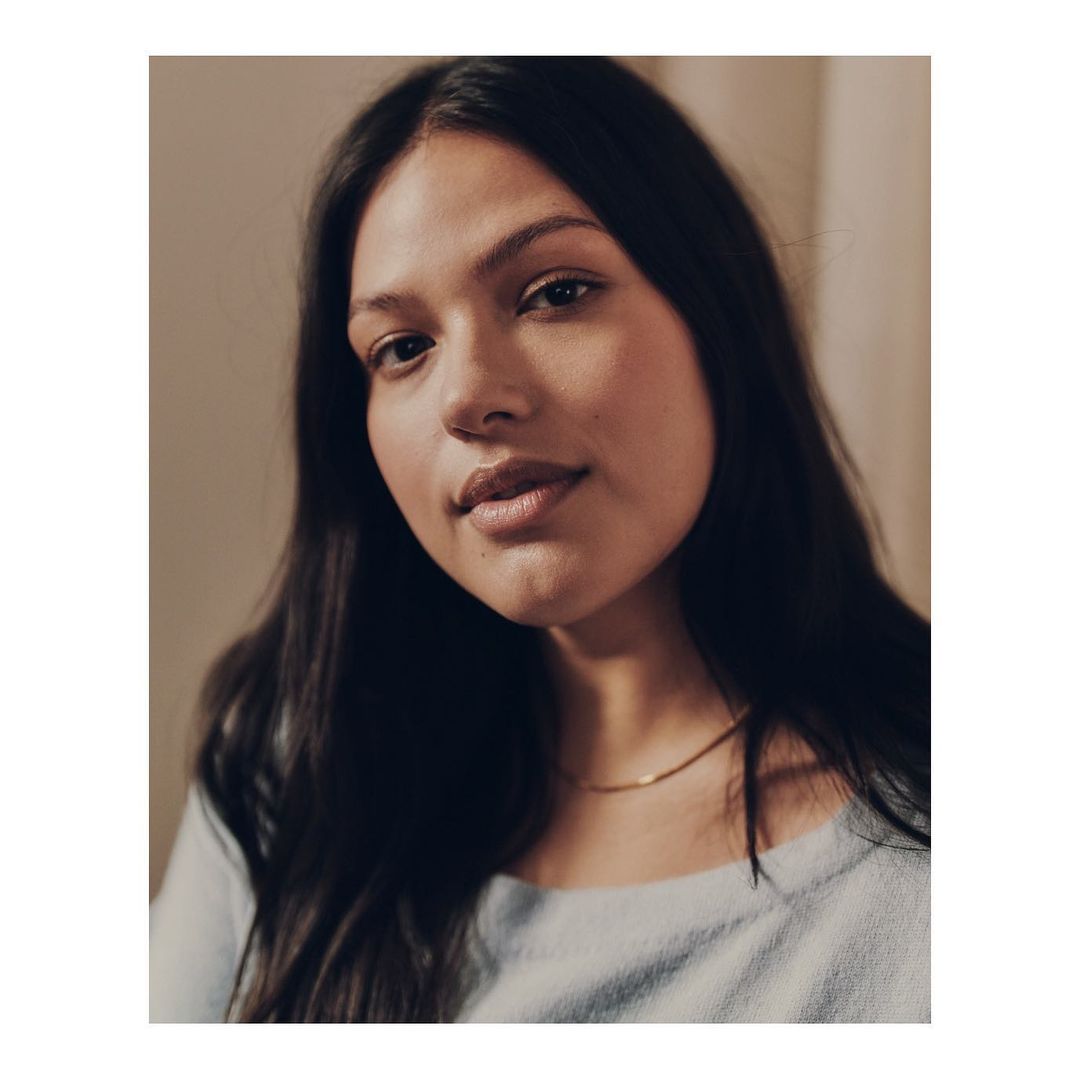 Close-up portrait of a woman with long dark hair, wearing a light blue top and a delicate gold necklace, with a neutral background.