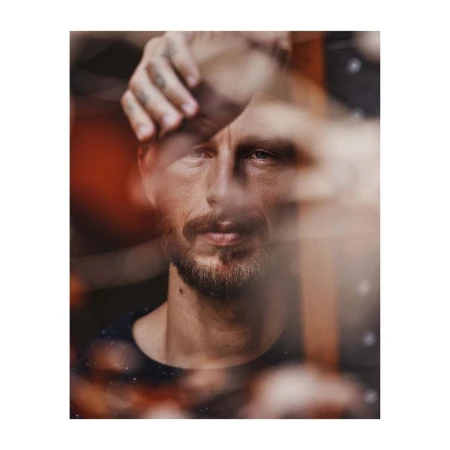 Close-up portrait of a man with facial hair framed through blurry foreground elements and hands