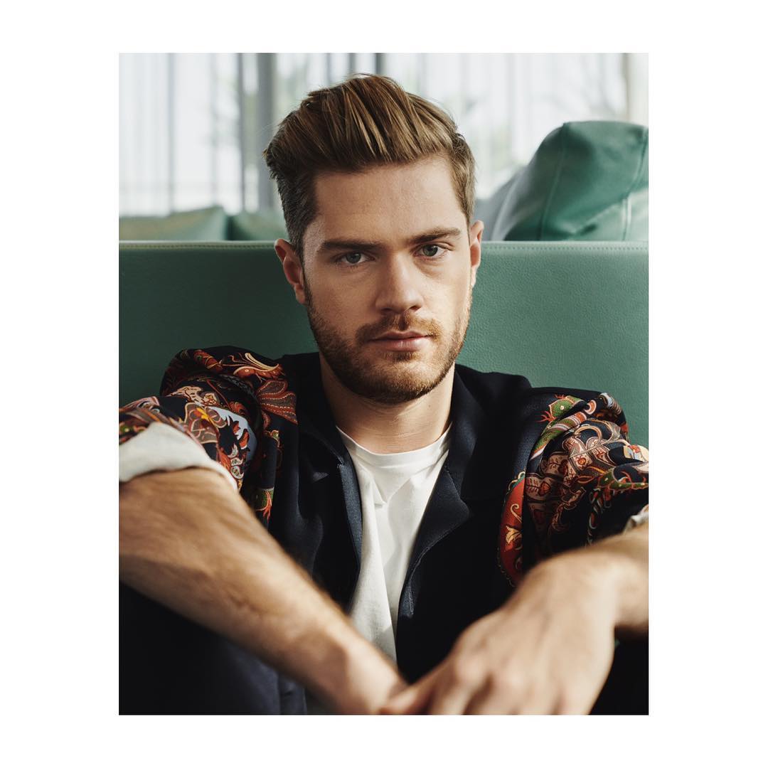 Portrait of a fair-skinned adult male with styled hair wearing a printed shirt with a blazer, looking directly at the camera indoors.