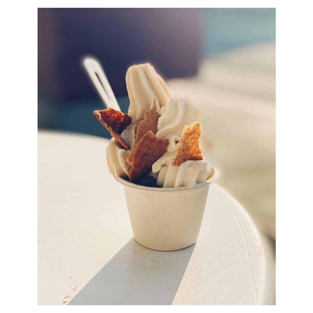 Close-up of a cup of soft serve ice cream with cracker pieces on top, set on a white table in an outdoor setting.