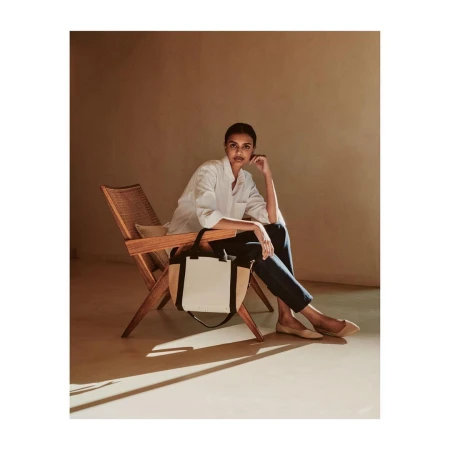 Woman in white shirt and dark trousers seated in a wooden rattan chair holding a two-tone canvas tote bag.