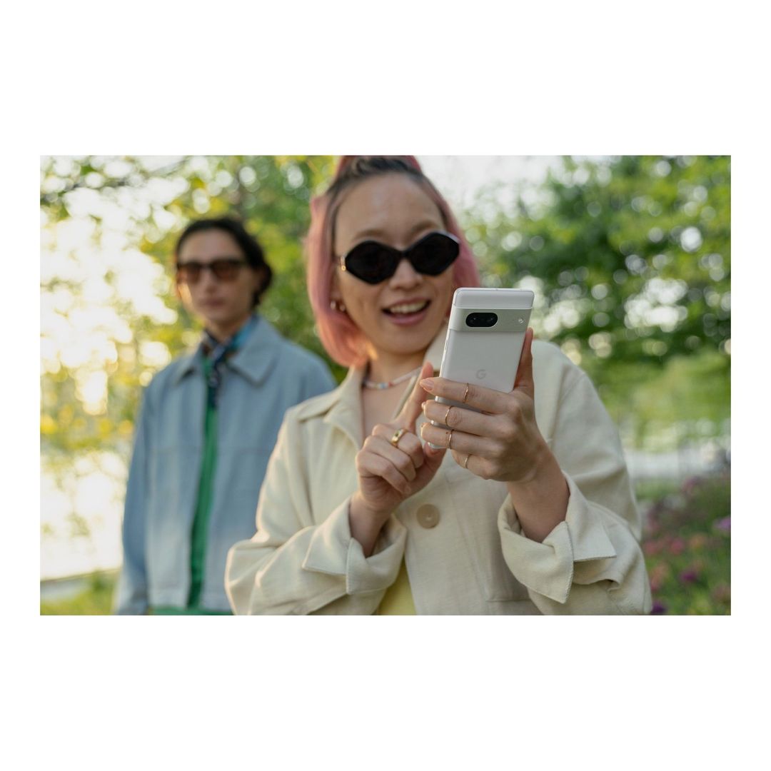 A smiling woman holds a Google Pixel phone outdoors with another person in the background, trees, and natural lighting.