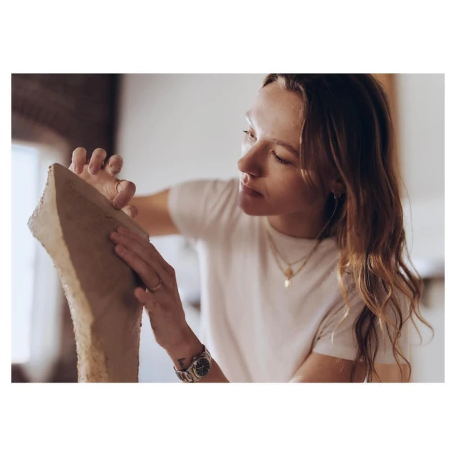 Woman sculpting a tall, narrow form with her hands. She wears a white t-shirt, and jewelry. Soft, natural light fills the space.
