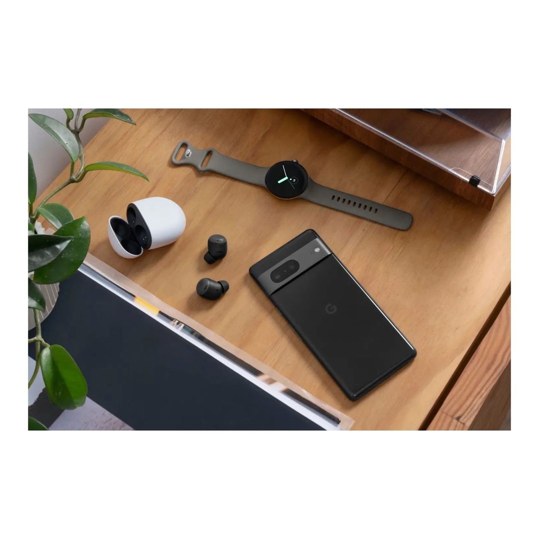 Arrangement of a black smartphone, gray smartwatch, and white wireless earbuds displayed on a wood-grain surface next to a green plant.