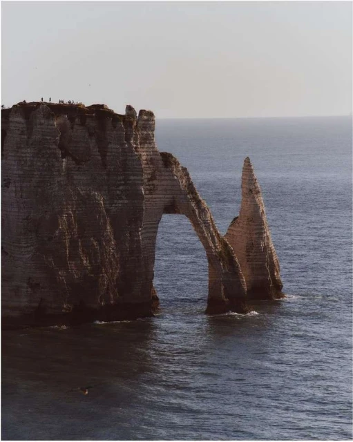 Dramatic seascape featuring the Étretat cliffs with its natural arch, bathed in soft light, with a calm ocean and a few figures on the cliffs.