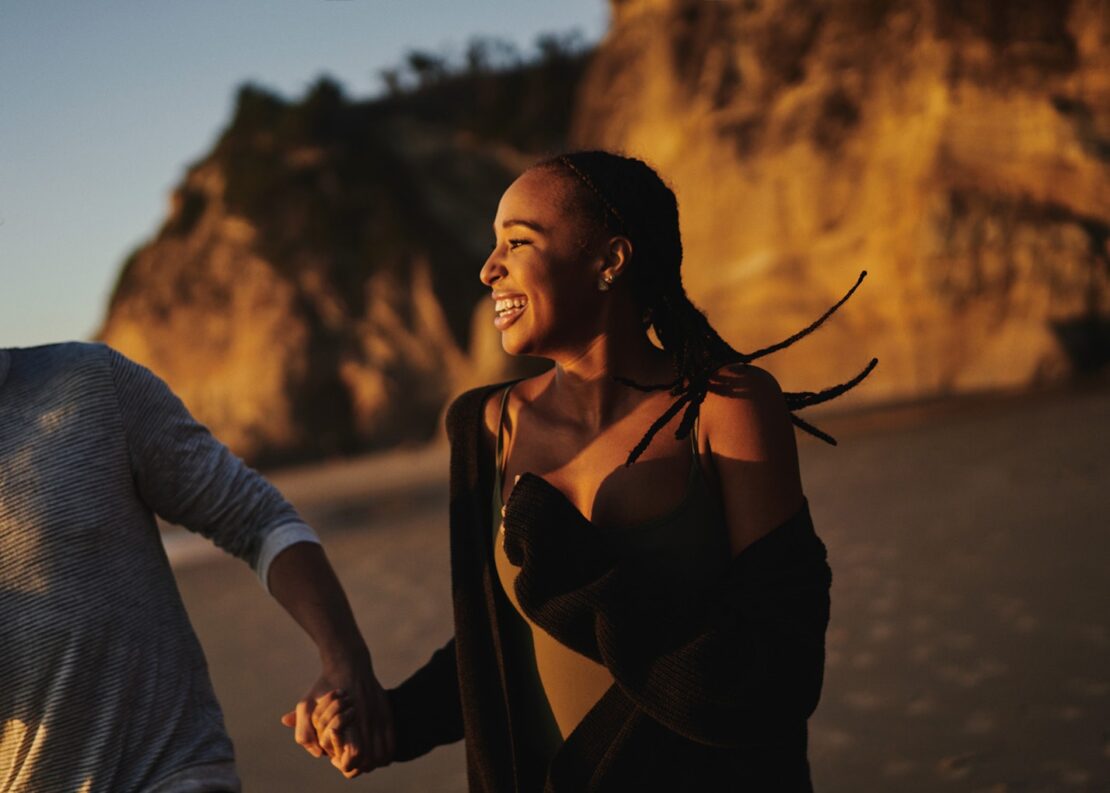 Smiling woman walking along a sandy beach at sunset holding hands with someone, captured by photographer Clayton Cotterell