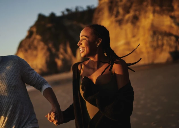 Smiling woman walking along a sandy beach at sunset holding hands with someone, captured by photographer Clayton Cotterell