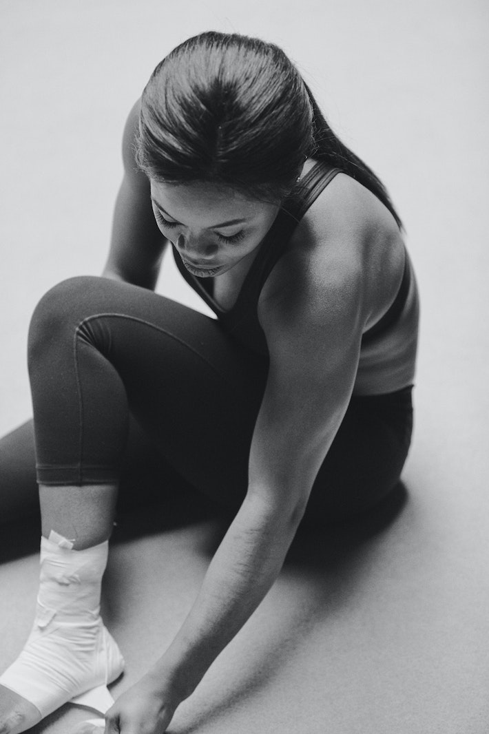 Athletic woman wraps her ankle with white bandage tape while seated on floor in fitness studio. Black and white image. 