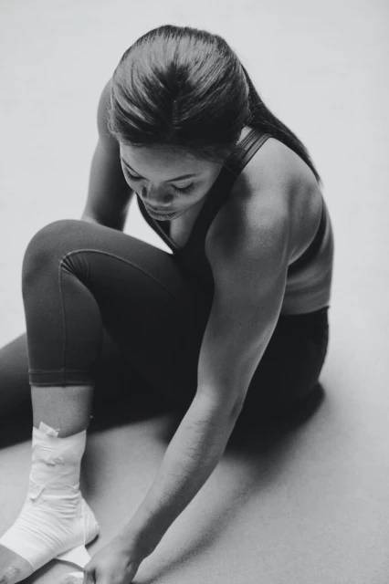 Athletic woman wraps her ankle with white bandage tape while seated on floor in fitness studio. Black and white image. 