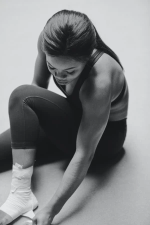 Athletic woman wraps her ankle with white bandage tape while seated on floor in fitness studio. Black and white image. 