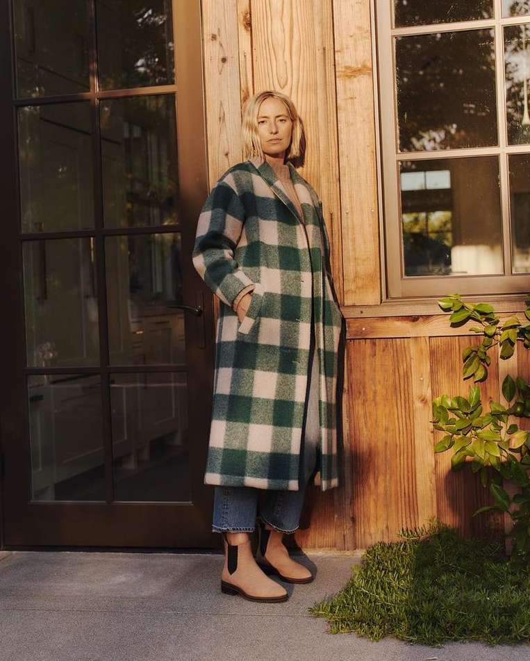 Adult woman in a green and white plaid coat standing in front of a wooden house with large windows and wearing tan-colored boots.