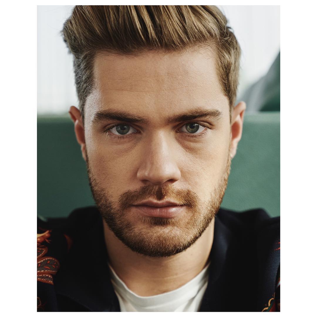 Close-up portrait of a handsome adult male with blond hair, a groomed beard, and green eyes, wearing a patterned top in soft, diffused lighting.