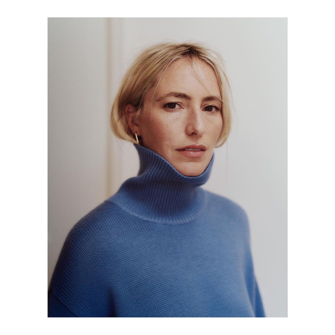 Portrait of a blonde woman wearing a blue turtleneck sweater against a plain white wall