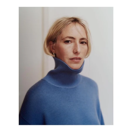 Portrait of a blonde woman wearing a blue turtleneck sweater against a plain white wall