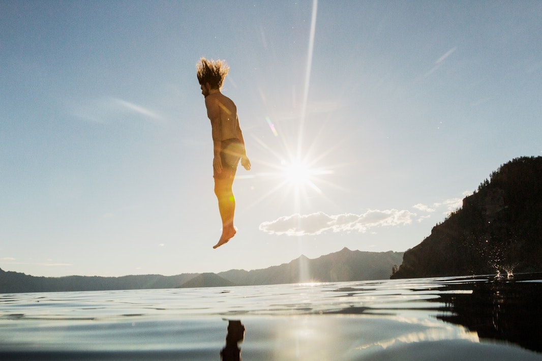 A man frozen mid-air jumping into water against a bright sunburst and distant mountainous horizon.