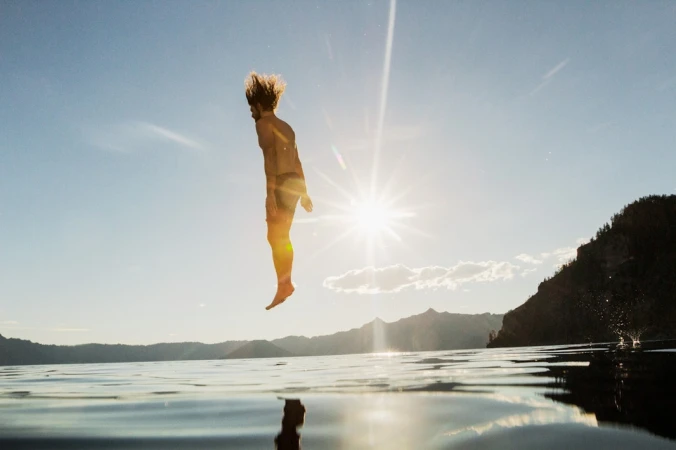 A man frozen mid-air jumping into water against a bright sunburst and distant mountainous horizon.