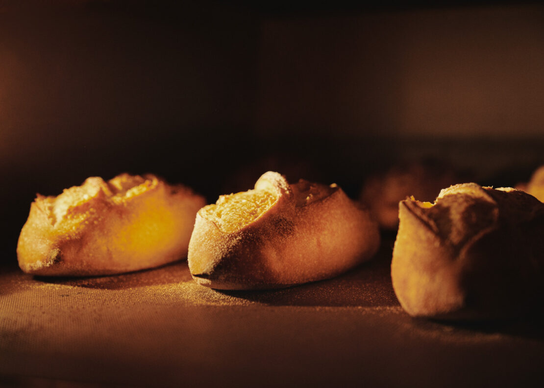 Close-up view of several golden-brown bread rolls baking in a warm-toned oven with a dark background, focus on texture and form.