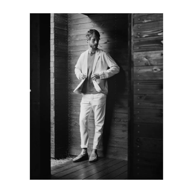 Monochrome portrait of adult male in light-colored jacket, t-shirt, and pants standing against a wooden door inside.
