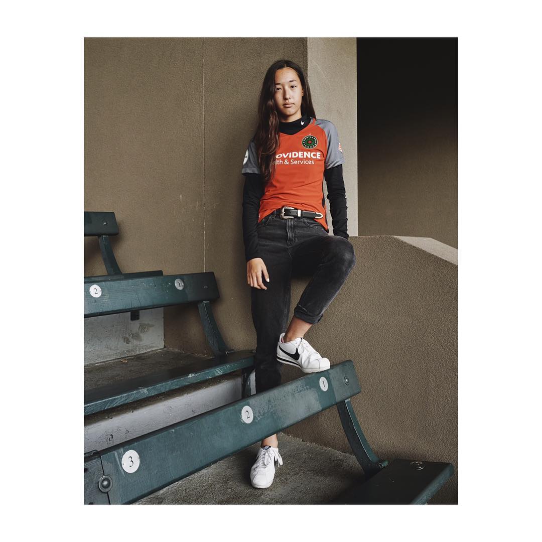Young woman in orange and black sportswear poses on teal concrete steps in an urban setting, showcasing sportswear and casual style.
