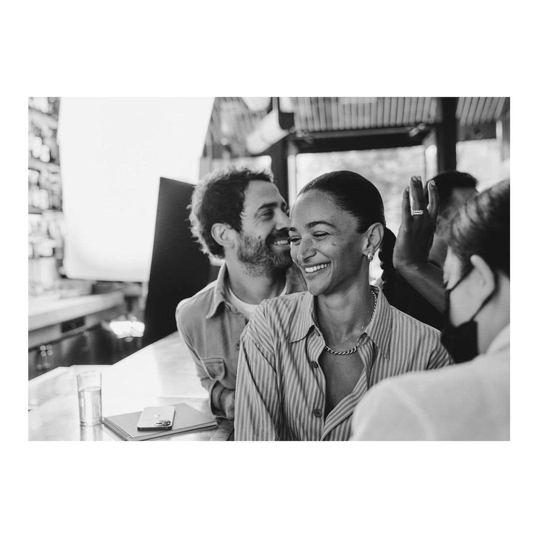 Black and white shot of a smiling adult woman and adult man at a bar, casually dressed with natural light, creating a candid, lifestyle moment.