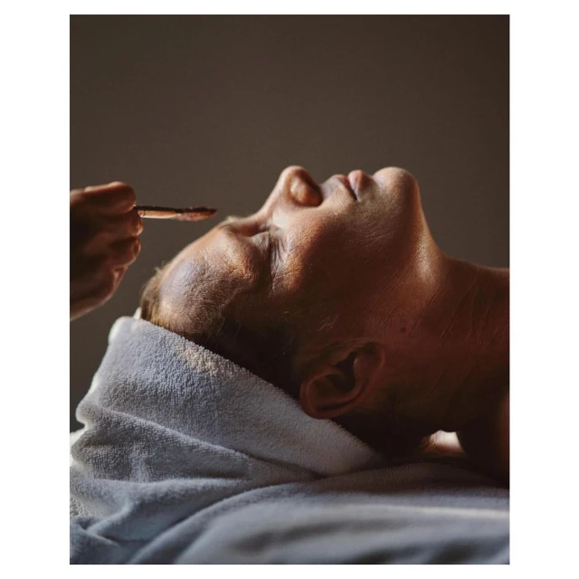 Adult man receives facial treatment with a towel around his neck, laying down with hands applying beauty product, inside a spa.