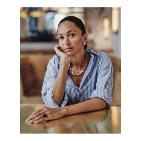 Woman in blue striped shirt leaning on a table with a gold chain necklace and rings in a cafe