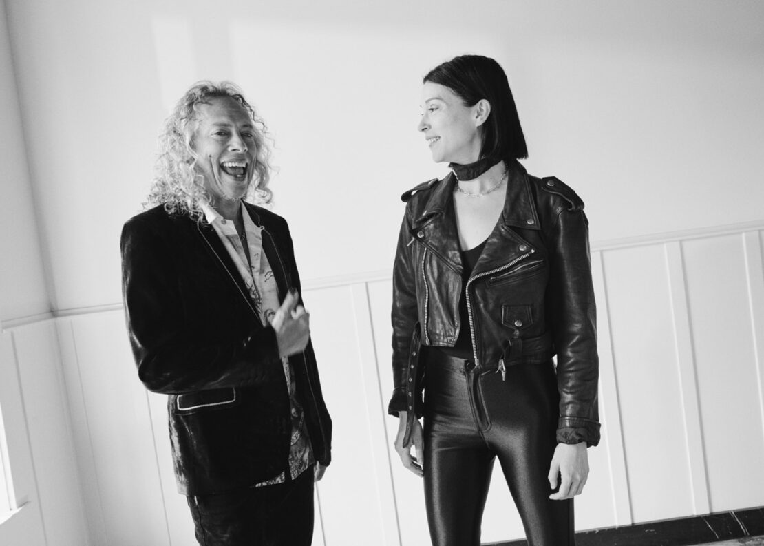 Black and white photo of Kirk Hammett in a blazer and a woman in a moto jacket, both smiling in a modern, indoor setting.