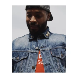 Portrait of a Black man wearing a denim jacket and a baseball cap backward against a plain light background