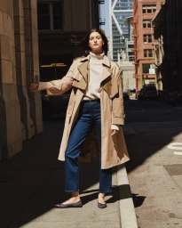 A woman wearing a beige trench coat, white turtleneck sweater, and blue jeans standing on a sunny city sidewalk.