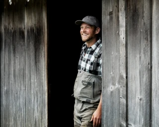 Smiling man in plaid shirt and grey waders peeking from wooden doorway