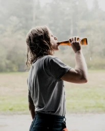 A person with wavy hair drinks from a glass bottle in an outdoor setting captured by Clayton Cotterell.