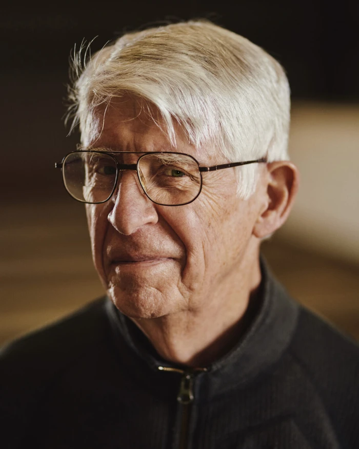 Close-up portrait of an elderly man with white hair and wire-rimmed glasses wearing a dark zip-up jacket