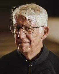 Close-up portrait of an elderly man with white hair and wire-rimmed glasses wearing a dark zip-up jacket