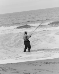 Person standing in the ocean surf on a sandy beach holding a fishing rod, captured in a monochrome photographic style.