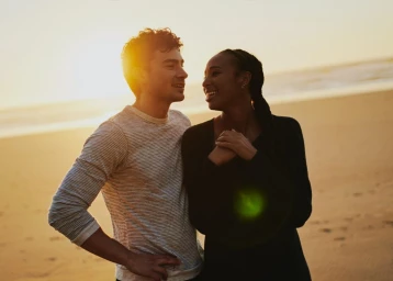 A smiling couple standing on a sandy beach at sunset, captured in warm golden hour light by photographer Clayton Cotterell.