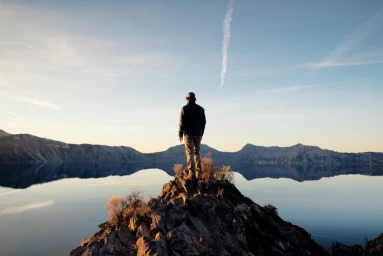 Man standing on a rocky outcrop overlooking a calm lake with distant mountain reflections under a clear sky.