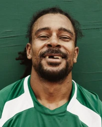 Close-up portrait of a Black man with dreadlocks wearing a green and white athletic jersey against a textured green background.