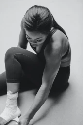 Black and white portrait of a woman wearing athletic wear and taping her ankle against a neutral seamless backdrop.