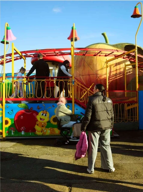Outdoor scene featuring a children's amusement ride with an apple theme, a woman holding a pink jacket, and several other people.