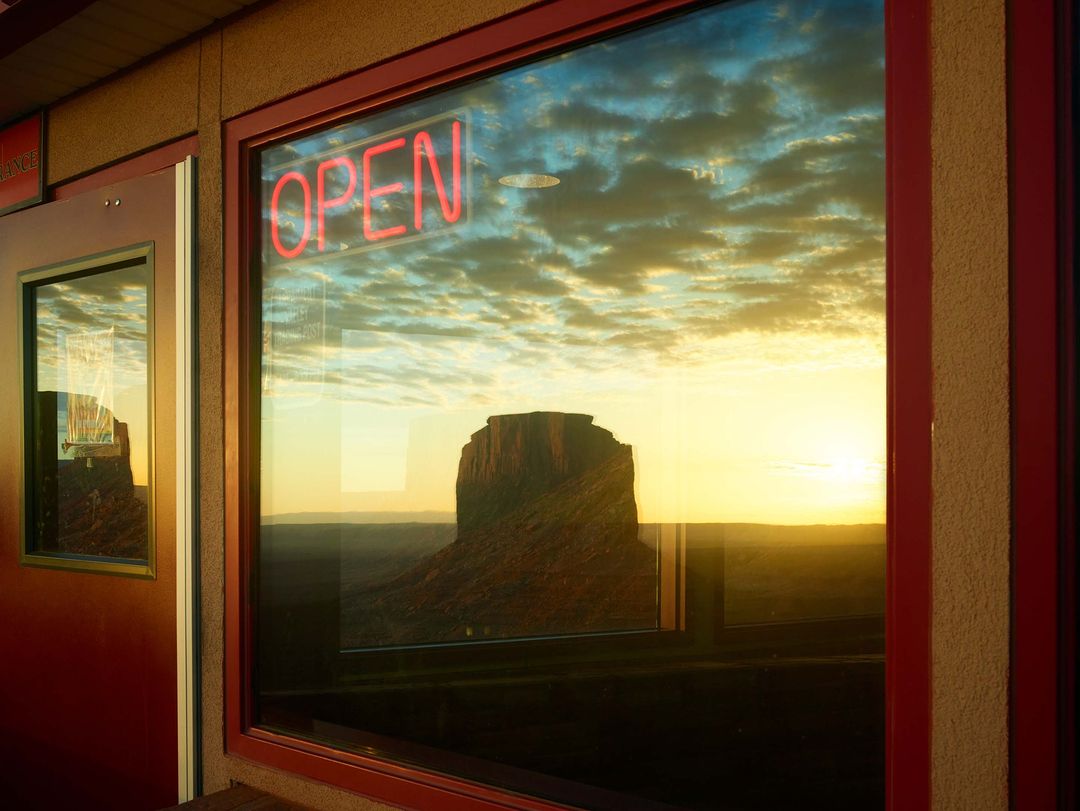 Monument Valley vista reflected in a window with a red neon 'OPEN' sign. Sunset sky and clouds add dramatic atmosphere.